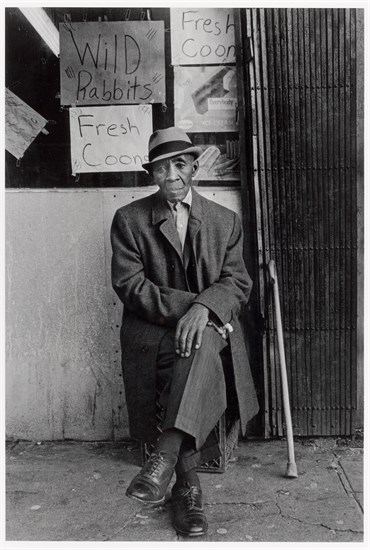 PFF203-Dawoud Bey, A Man at 116th and Lenox Avenue, 1976. Black and white photograph of an older black man in suit and hat seated within what appears to be a market with handwritten signs in the background.